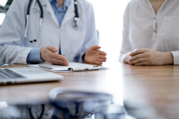 Doctor and patient discussing something while sitting near each other at the wooden desk in clinic, close up of hands. Medicine concept