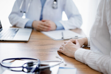 Doctor and patient sitting and discussing something at wooden table while using a laptop computer. Focus is on patient's hands. Medicine concept