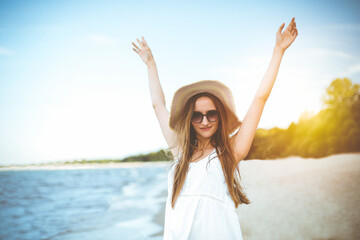 Fototapeta na wymiar Happy smiling woman in free happiness bliss on ocean beach standing with a hat, sunglasses, and rasing hands. Portrait of a multicultural female model in white summer dress enjoying nature