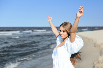Happy, beautiful woman on the ocean beach standing in a white summer dress, raising hands