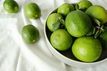 vários limões verdes dentro de prato cinza sobre mesa com tecido branco com folha de hortelã
