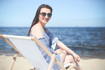 Happy young brunette woman relaxing on a wooden deck chair at the ocean beach while looking at camera, smiling, and wearing fashion sunglasses. The enjoying vacation concept