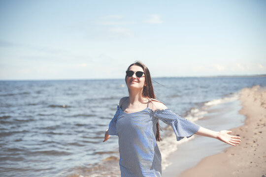 Happy smiling woman in free bliss on ocean beach standing with open hands. Portrait of a brunette female model in summer dress enjoying nature during travel holidays vacation outdoors