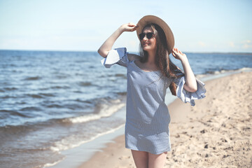 Happy young brunette woman standing on the ocean beach while smiling, and wearing fashion hat, sunglasses. The enjoying vacation concept