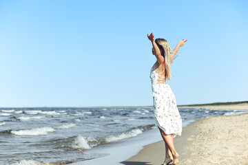 Happy blonde beautiful woman on the ocean beach standing in a white summer dress, open arms