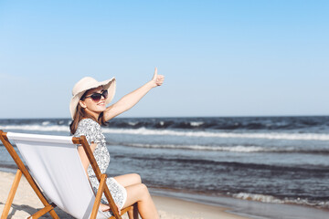 Fototapeta na wymiar Happy brunette woman sitting on a wooden deck chair at the ocean beach while showing Thumb up