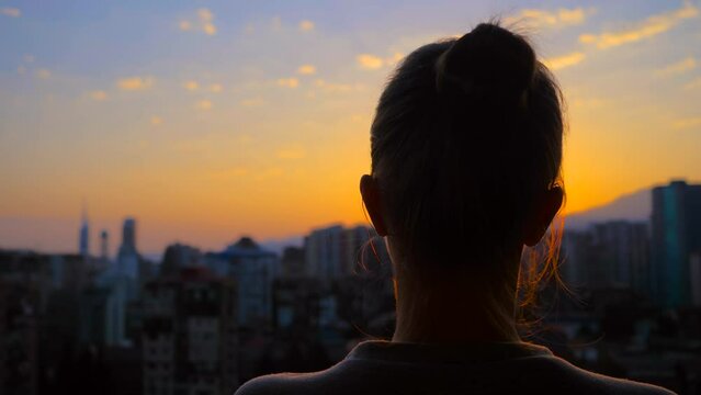 Back view: woman silhouette is standing on the balcony and looking at the sunset, sunrise sky over the city - close up, sun lens flares. Lonely, urban, dramatic and freedom concept
