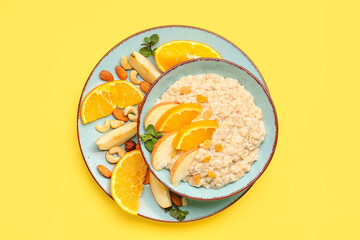 Bowl with tasty oatmeal, orange, apple and nuts on yellow background
