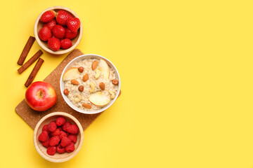 Bowl with tasty oatmeal, nuts, ripe berries, apple and cinnamon on yellow background