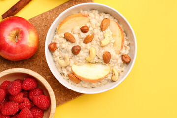 Bowl with tasty oatmeal, nuts, raspberries and apple on yellow background
