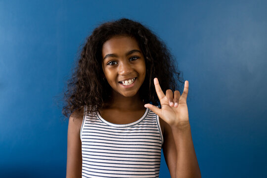 Portrait of happy biracial girl using sign language with hand on blue background - Powered by Adobe