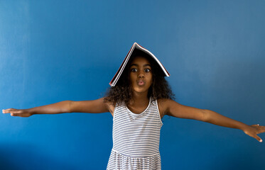 Portrait of biracial schoolgirl with arms outstretched and notebook on head, on blue background