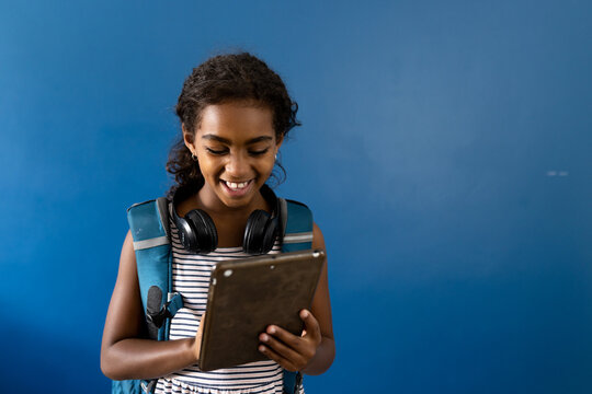 Happy biracial schoolgirl with headphones using tablet, smiling on blue background with copy space - Powered by Adobe