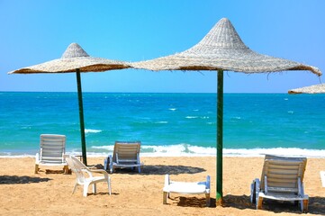 A deserted beach in the Sahara with empty white chairs and umbrellas on the sand and a beautiful blue horizon where the sea water meets the blue sky.