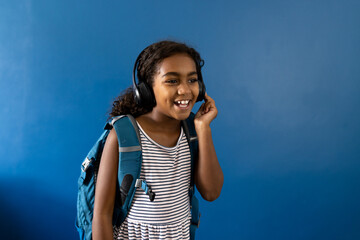 Happy biracial schoolgirl wearing headphones listening to music on blue background with copy space