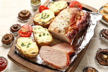 Wooden board with tasty baked meat, potatoes, tomatoes and mushrooms on light tile background, closeup