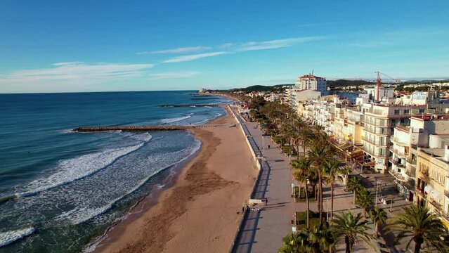 Drone flight back over in along coast beautiful beaches surrounded by Palm trees in Sitges. Mediterranean landscape of beach and hotels. Gentle sea waves quietly roll onto shore. Film intro. 4K video