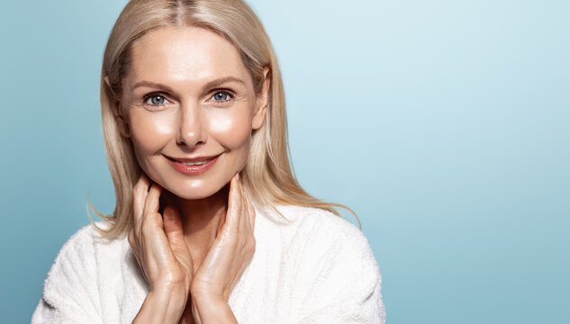 Smiling 50s middle aged mature older woman applying facial cream on face looking at camera isolated on blue background. Anti age healthy dry skin care beauty therapy concept, old skincare treatment