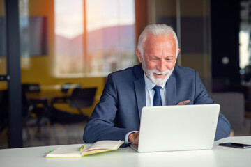 Handsome smiling senior man while sitting at his cozy workplace with laptop at home, retired male chatting with friends in social media,