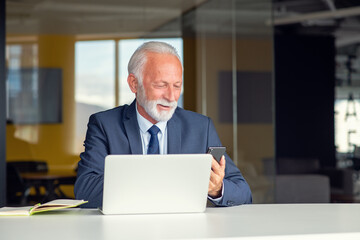 Handsome smiling senior man while sitting at his cozy workplace with laptop at home, retired male chatting with friends in social media,