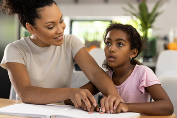 Smiling biracial mother sitting at kitchen table helping her blind daughter to read braille