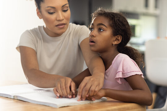 Biracial mother sitting at table helping her blind daughter to read braille