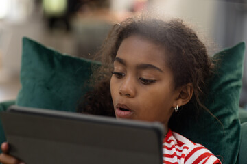 Happy biracial girl lying on couch making video call using tablet