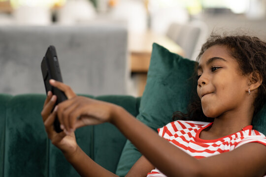 Happy biracial girl relaxing on couch, smiling and using smartphone