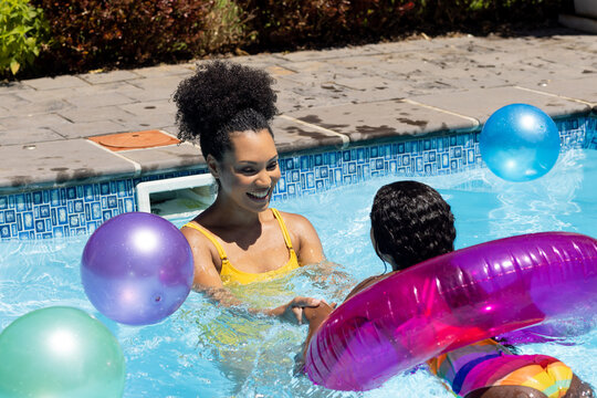 Happy biracial mother and daughter playing with inflatable in sunny outdoor swimming pool