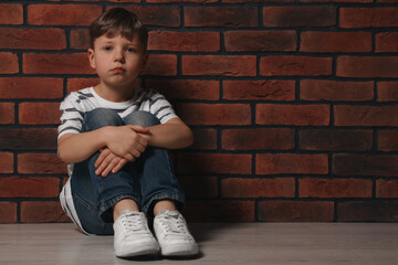 Little boy sitting on floor near brick wall, space for text. Children's bullying