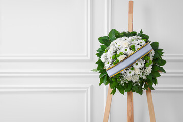 Funeral wreath of flowers with ribbon on wooden stand near white wall indoors. Space for text