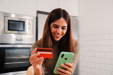 Young caucasian female with white perfect teeth smiling and using a cell phone app to purchase and pay with a credit card sitting at home kitchen. Woman spending money on online shopping on internet