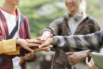 A group of diverse, multiracial young people are shown happily stacking their hands together while standing outside a building. This represents a sense of unity, fun,relationship and youth culture.