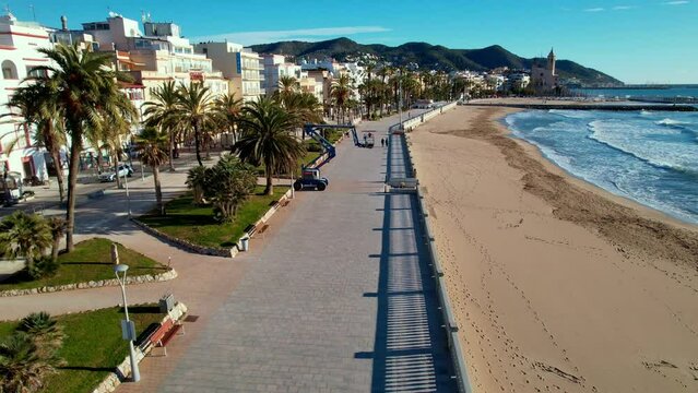 Panoramic aerial drone view of coastal village, Sant Bartomeu i Santa Tecla de Sitges in Sitges. People are walking along embankment with classic white balustrade and empty terrace overlooking sea. 4K