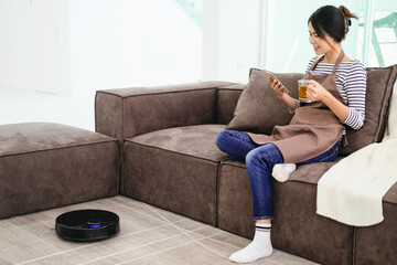 Black robotic vacuum cleaner cleaning the floor while woman sitting on sofa and relax.Young woman...