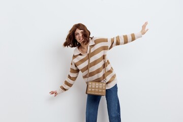 a happy, joyful woman stands on a white background in a stylish striped sweater and blue jeans, funny leaning to the side and raising her arms, wearing a bag over her shoulder