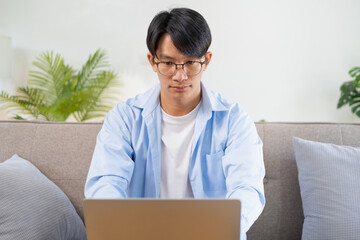 Handsome young businessman in eyewear working with computer remotely, sitting at wooden table in living room. Pleasant happy man communicating in social network, searching information online..