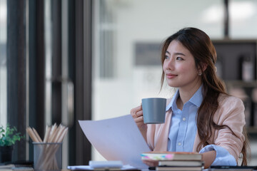 An attractive businesswoman holding a cup of hot drink and looking away with a smile while sitting in an office.