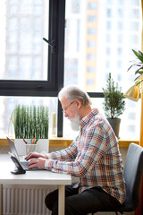 Serious elderly man using laptop checking email news online sitting in living room, searching for friends in internet social networks or working on computer, writing blog or watching webinar, at home