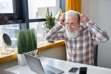 senior 70s man sit relax in cozy room browsing internet on modern laptop and listen to music using...