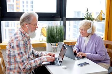 Senior Caucasian People Using Laptop Working Or Studying At Home, Websurfing In Cozy Room. Older Couple Sit Opposite Of Each Other Browsing Internet On Laptop Reading Online News On Weekend