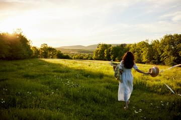 a woman in a light dress stands in a field during sunset with a wicker basket, a hat and a plaid in her hands
