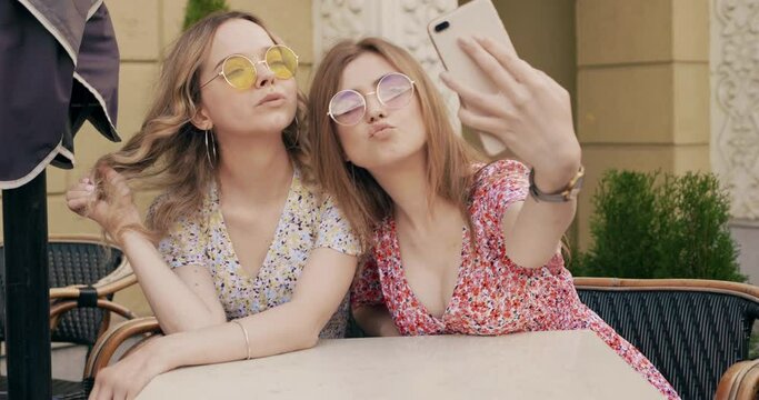 Two young beautiful smiling hipster girls in trendy summer sundress. Carefree women chatting in veranda cafe on the street background. Positive models having fun and taking selfie on smartphone