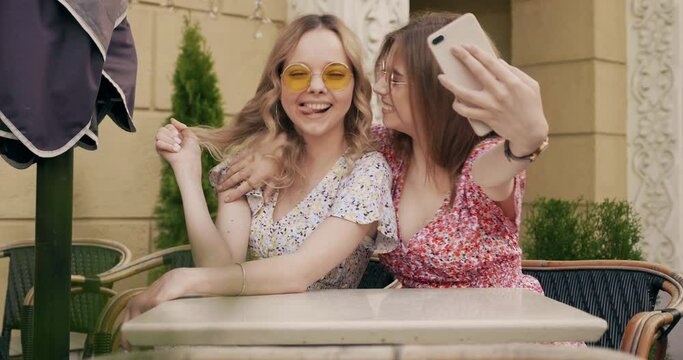 Two young beautiful smiling hipster girls in trendy summer sundress. Carefree women chatting in veranda cafe on the street background. Positive models having fun and taking selfie on smartphone