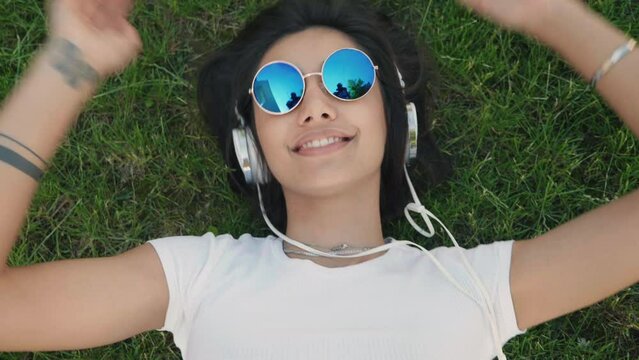 Fashion portrait of young stylish hipster woman lying on the grass in the park. Girl wears trendy outfit. Smiling model enjoy her weekends. Female listening music via headphones and singing. Top view