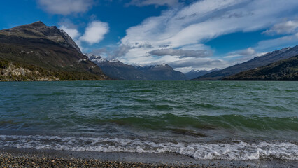 The lake is surrounded by picturesque mountains. Ripples on the emerald water. The waves are foaming on the coastal pebbles. Blue sky with clouds. Tierra del Fuego National Park. Lago Roca. Argentina.