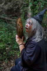 old witch with white hair and a black hat kissing her broom with a forest in the background