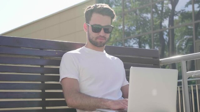 Handsome smiling stylish hipster lambersexual model. Fashion man sitting on the bench in the street and using laptop computer, writing text. Concept of co-working startup. Technology and communication