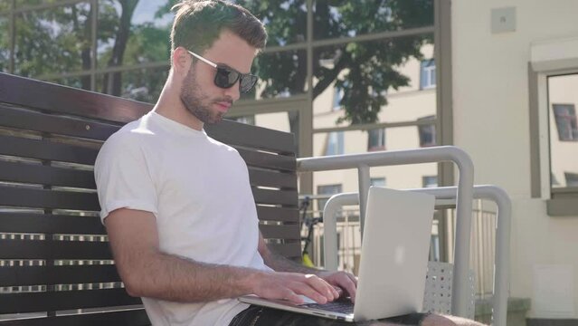 Handsome smiling stylish hipster lambersexual model. Fashion man sitting on the bench in the street and using laptop computer, writing text. Concept of co-working startup. Technology and communication