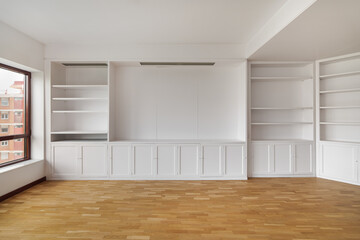 Fototapeta premium Empty built-in white cabinet on wooden laminate floor with big window in empty bright airy room in housewarming. Concept of housewarming and convenient storage